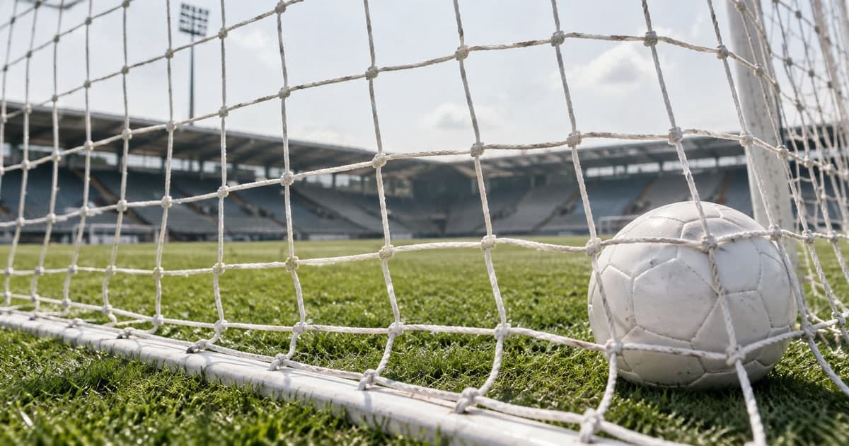 흐린 낮 하늘 아래 축구 골대 그물 안쪽 잔디 위에 흰 축구공 하나가 놓여 있고, 그물 너머로 초록 경기장과 빈 관중석, 조명탑이 흐릿하게 보인다.